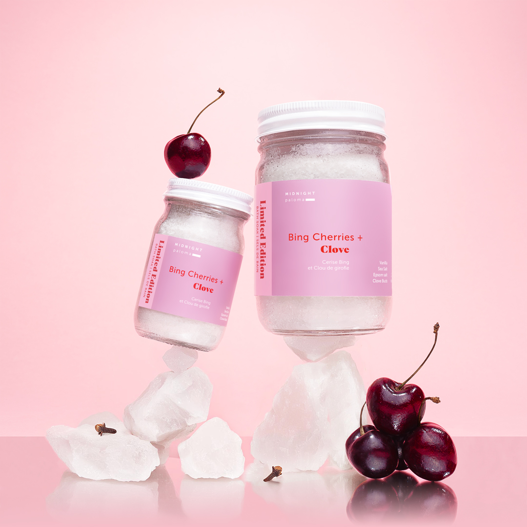Limited edition Bing Cherries + Clove Bath Soak in glass jars, styled with fresh cherries and cloves on a pink background. Creative direction by Danielle Gracioso.