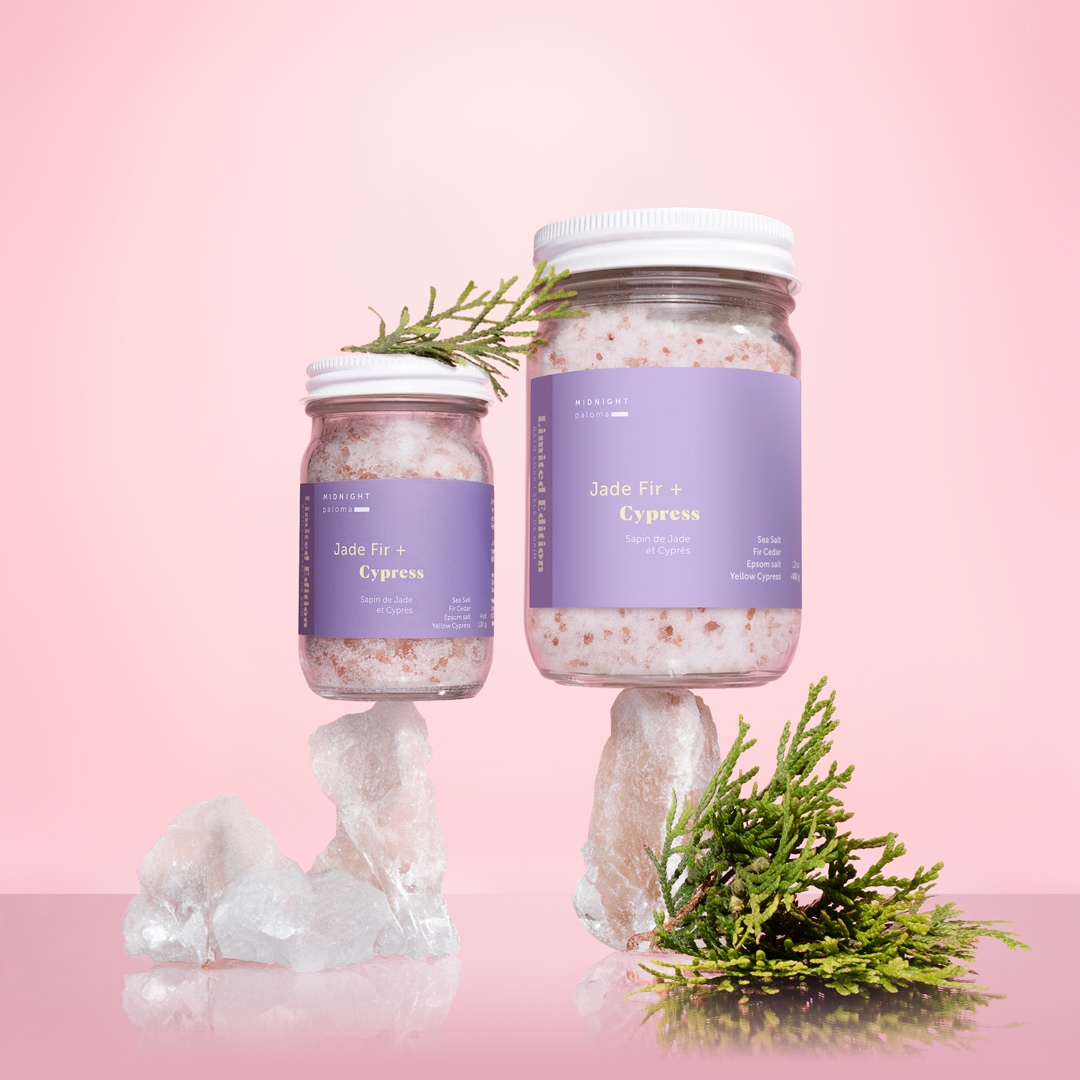 Jade Fir + Cypress Bath Soak in two jar sizes with fir sprigs and bath salts on a pink background. Visual identity and photo concept by Danielle Gracioso.